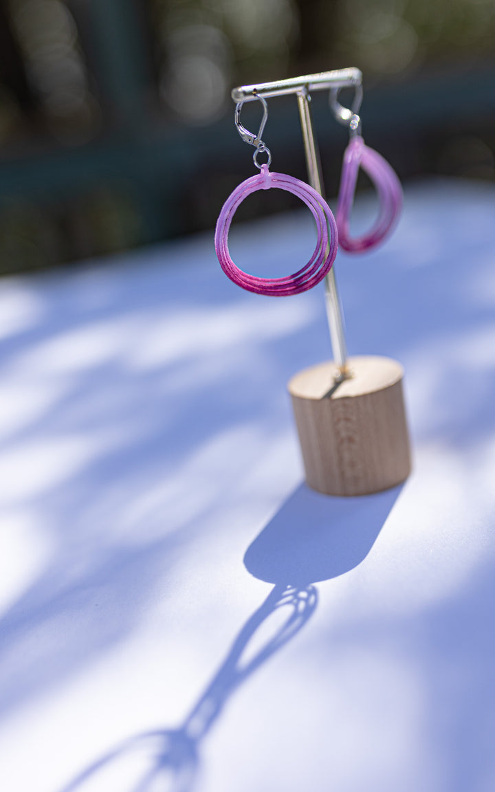 Boucles d’oreilles rondes roses, suspendues à un présentoir sous la lumière douce du matin. Teintes ombrées appliquées à la main sur nylon recyclé. Bijou 3D ultraléger fabriqué au Québec par Vox & Oz.
