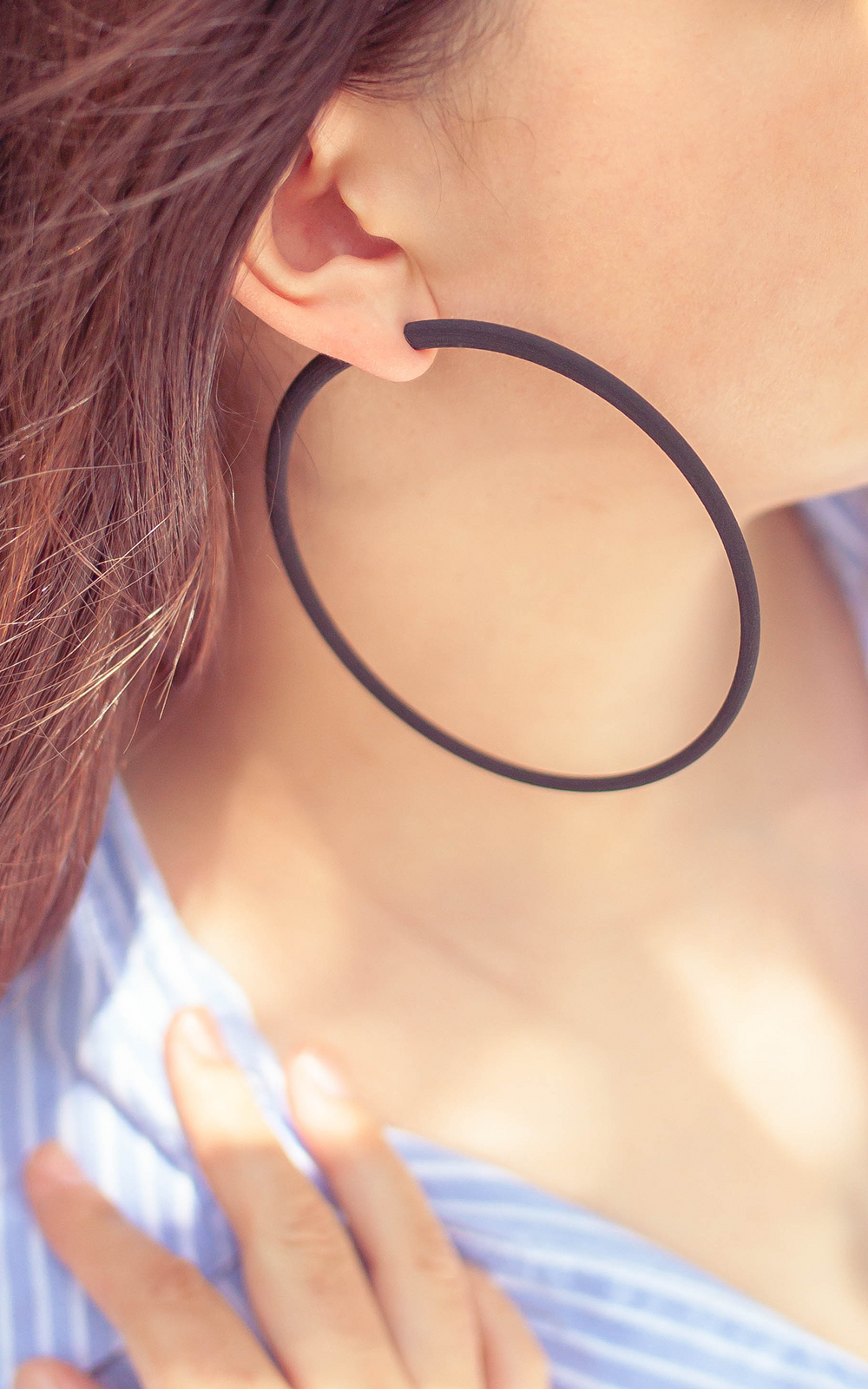 Les anneaux Atlas 10 cm De grands anneaux ultralégers ! Un poids plume ! Confort et design aux lignes minimalistes au tracé épais. Porter par jeune femme cheveux bruns. Saison été. Couleur noir.